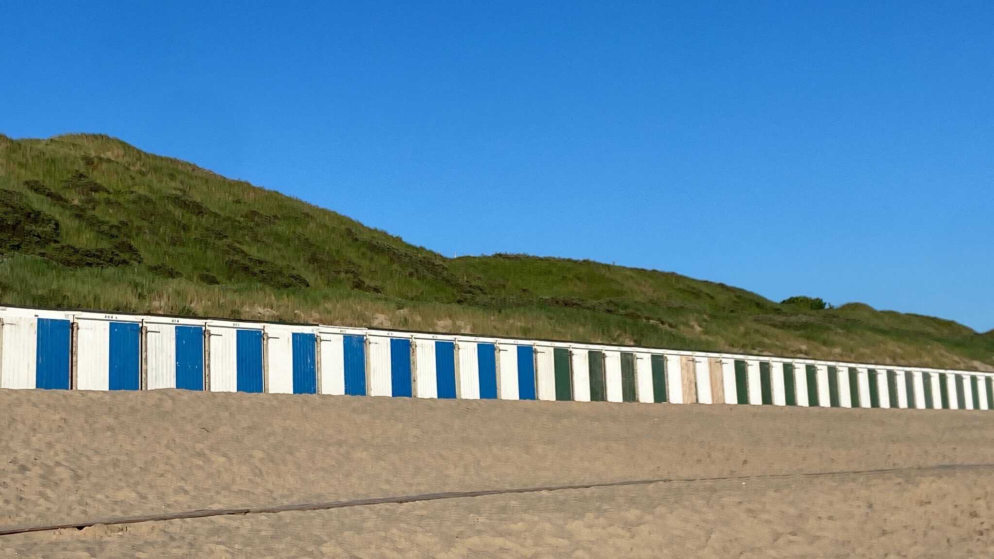 Image description: row of tiny white beach garages with blue green and beige doors on sandy surface, they kind of look like a piano keyboard, behind them a grassy hill and cloudless sky