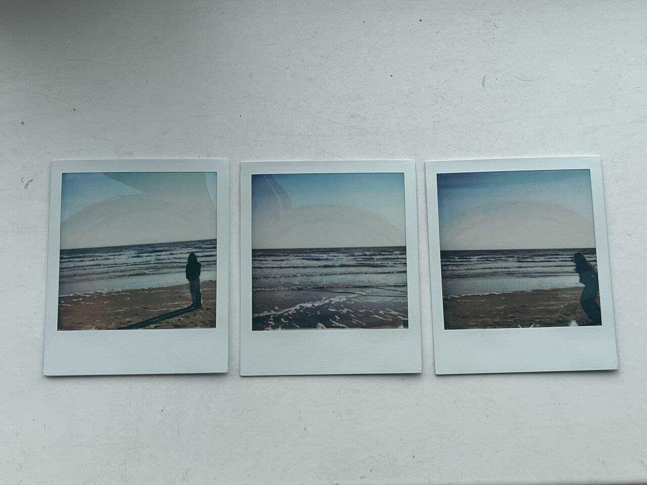 Three polaroid placed on white surface, all looking into the distance of the sea - a bit of the sandy beach and the waves that are washing over it. the sky is overexposed into whiteness with a denim aureola, with a person standing in the left polaroid and one running into the frame in the one on the right. the horizon lines of the photographs are lined up, even though the angles at which they have captured the waters differ slightly